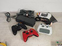 Three Xbox 360 wireless controllers (black, red, white) laid out with cords and a white chatpad keyboard attached to one controller, power brick and AV cables in background.