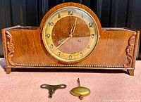 Full front view of the wood mantle clock with brass bezel round face showing time 12:02 and visible Arabic numerals, flanked by carved floral details on each side and beaded trim on the lower front edge with winding key and pendulum placed before it.