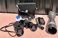 Wide photo showing a portable binocular in carrying case, full-sized binoculars, field scope with lens, and accessories on a flat surface outside.