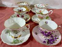 Six pairs of fine bone china teacups and saucers arranged on a pink cloth showing various floral patterns and gold trim.
