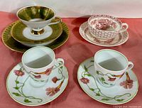 Four sets of fine bone china cups and saucers arranged on a red cloth, showing intricate designs including gold detailing, floral patterns in red and pink, and green vines.