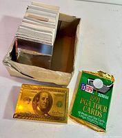 View of large box containing a thick stack of standard sized vintage sports trading cards including shiny reflective cards visible front and center. Next to it is a lenticular gold playing cards deck with Benjamin Franklin gold bill design and a 1991 Pro Set PGA Tour Golf Cards pack in green and gold packaging.