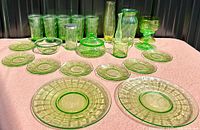 Group display of glassware including tumblers, plates, covered dish, vase, and bowl, arranged on pink cloth