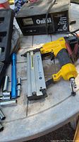 Yellow pneumatic air nailer, Omnia battery charger, blue flashlight, and orange clamp on white table.