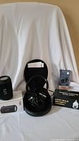Black Sony over-ear headphones inside a hard carrying case with cables, wireless earbuds in charging case with packaging, and boxed light bulb security camera on white backdrop.