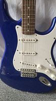 Close-up of the blue guitar body showing white pickguard and three single-coil pickups with control knobs and bridge.