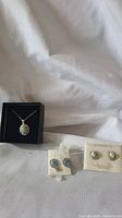 Photo showing all jewelry items on white fabric background: one sterling silver necklace with a green oval gemstone pendant in a black box, and two pairs of earrings with blue gemstones and pearl-like centers on white cards.