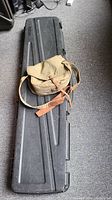 Two items: a long black hard weaponry case and a vintage tan canvas military bag with leather straps, both placed on a carpeted floor.