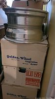 Side view of a tire rim showing polished silver metal surface, stacked on top of boxes labeled Golden Wheel Corporation and Enkei, made in Japan.