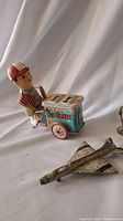 Side view of metal wind-up ice cream seller toy pushing a blue ice cream cart with red wheels; metallic Concord airplane toy model shown partially in foreground.