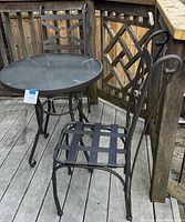3 piece outdoor bistro set on a wooden deck. Round glass top table with two black metal chairs featuring strap seats and decorative backs. Minor rust spots on chair legs visible.