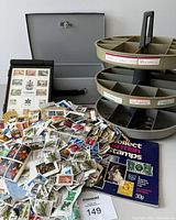 Overall view of the lot showing framed Canadian Centennial stamp collection, loose stamps, British stamp booklet, plastic carousel storage with labeled compartments, and gray metal storage box.