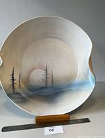 Front view of ceramic plate showing landscape with sun and three pine trees in soft natural tones. Plate on wooden stand with ruler for size reference.