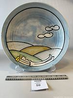 Front view of the round glazed ceramic plate with Cubist-style hand painted design including a reclining figure and hills under three clouds, pastel colors on blue background