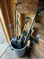 Various garden and snow shovels, rakes and other tools packed standing in a large gray garbage pail inside a wooden shed