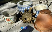 Five garden items photographed on outdoor deck: four ceramic pots (colorful floral, blue-and-white patterns, and terracotta), Mayan terracotta sun sculpture (broken), small ruler for scale.