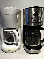 Two coffee makers, white Philips 12-cup and black Insignia 12-cup programmable drip coffee makers, side by side