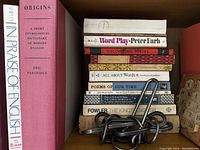 Shelf showing a stack of language and word books, including titles like "Origins," "Word Play," "All About Words," and other poetry and language related books, with a metal puzzle resting on top of the stack.