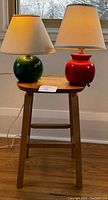 View of two ceramic lamps, one red and one green, sitting on a wooden stool in front of a window.