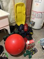 Photo showing red exercise ball, green dumbbells, boxed Pilates starter kit, camping pillows and sleeping bags, duffle bags, and yellow plastic sled against basement wall.