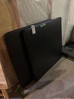 Two black padded card tables leaning against a basement wall, stacked one in front of the other, showing the full top surfaces and edges.