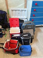 Photo showing a variety of cooler bags and a large white Coleman cube cooler placed on the floor in a basement setting, highlighting the different sizes and colors.