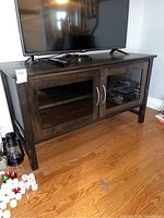 Front view of the wooden credenza with dark finish, glass double doors with handles, placed on hardwood floor with a TV and some small items on the top and sides.