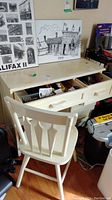 Front view of the cream painted wooden desk with multiple drawers open and cream painted wooden chair in front. Desk top and chair show paint wear and stains.
