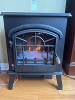 Front view of black metal Kamado electric fireplace space heater with glowing logs inside, showing the glass door with windowpane design and decorative handle.