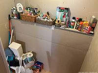 View of various cleaning supplies and household items organized on a narrow shelf and floor beneath, including fire extinguishers, tape, cleaning sprays, and dustpans.