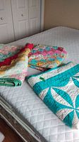 Three folded quilts on mattress showing blue and white, bright multicolor, and green border reversible patterns.