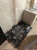 Photo of open black plastic tool case placed on floor showing tools arranged inside including wrenches, ratchets, sockets, pliers, screwdriver bits, hammer, level and measuring tape.