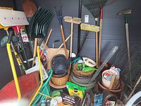 Photo showing red metal wheelbarrow, snow shovels, various garden rakes, garden hoe, watering can, flower pots, and bags of potting mix with additional garden tools and accessories stacked inside a shed
