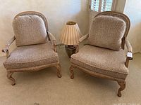 Two oversized upholstered armchairs in patterned beige fabric and wooden frames with cabriole legs, alongside a pleated shade table lamp.