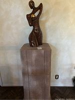 Full view showing the abstract woman sculpture displayed on a tall stand against a cream textured wall.