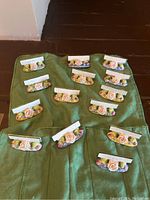 Set of 14 antique porcelain place-card holders arranged on green cloth, each with floral decoration and space for cards with names.