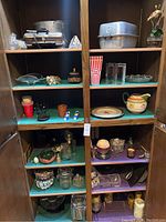 Full view of two cabinet shelves with household items including metal pans, glassware, pottery, and decorative pieces.