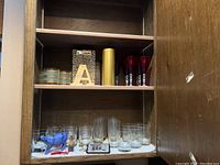 Upper shelf of wooden cabinet with small blue and gold striped containers, leopard print and gold letter 'A', large gold cylindrical container, and several colored wine glasses in red and purple.