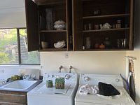 View of laundry room showing upper wooden cabinets with dishes, two laundry sinks, and laundry machines with items placed on top