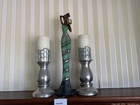 Photo showing two silver-tone metal candle holders with white candles and a tall female figurine in a green and black dress.