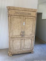 Front view of large beige armoire showing full height, ornate carved details and leaf motif on upper door panel.