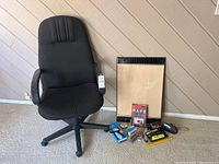 Black fabric office chair, electric heater, stapler, tape dispenser, batteries, plug adapter, and book on carpeted floor against wood panel wall.
