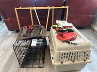 Photo showing 2 dog carriers: a large beige plastic crate and a smaller black wire metal crate, along with a red dog leash and several collars and harnesses on top of the plastic crate.