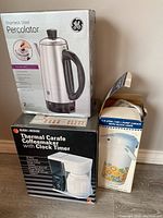 Photo of boxed General Electric stainless steel percolator, Black & Decker thermal carafe coffeemaker with clock timer box, and 1.9L pump carafe with floral design in box.