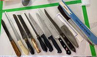 Full view of 9 knives of various handle and blade types laid out in a row on a surface with green tape edges