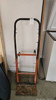 Front view of the 4-foot tall dolly, showing the black handle, orange frame, and rusty metal platform base.