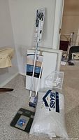 Two white laundry drying racks leaning against the wall, jumbo pillow in original Sears packaging, telescoping bulb changer, rectangular glass scale with black frame on floor