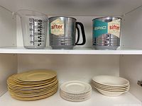 Two metal Epic flour sifters, 5-cup measuring cup, stack of yellow/golden plates and smaller white plates