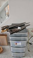Three vintage hand saws placed on top of a plastic drawer unit. The saws have wooden handles with different colors and the blades show wear.