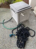 Photo showing gray plastic hose winding box with hose inside and black expandable garden hose with attached spray nozzle.
